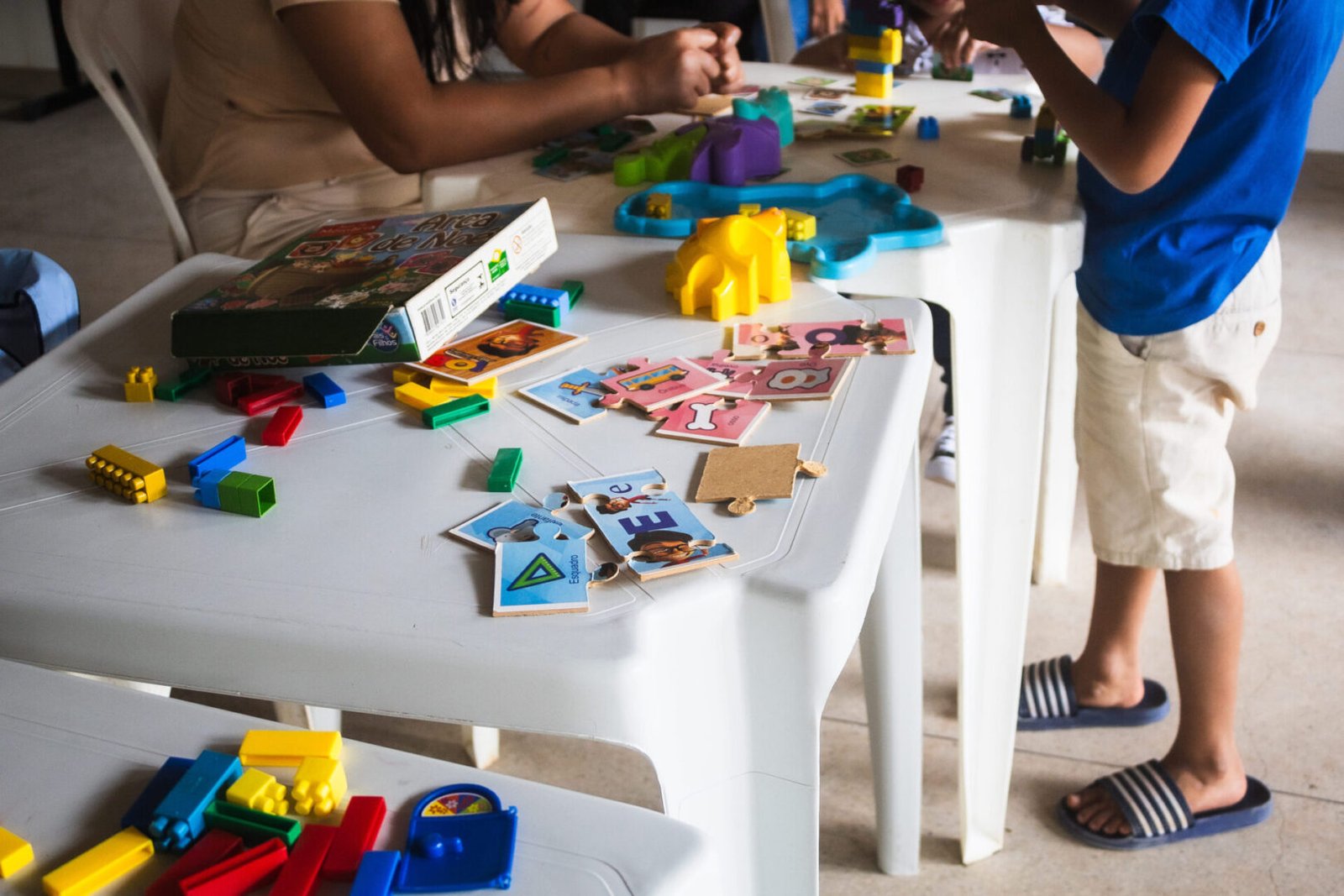 Mesa de plático branca com brinquedos coloridos em cima e uma criança perto. 